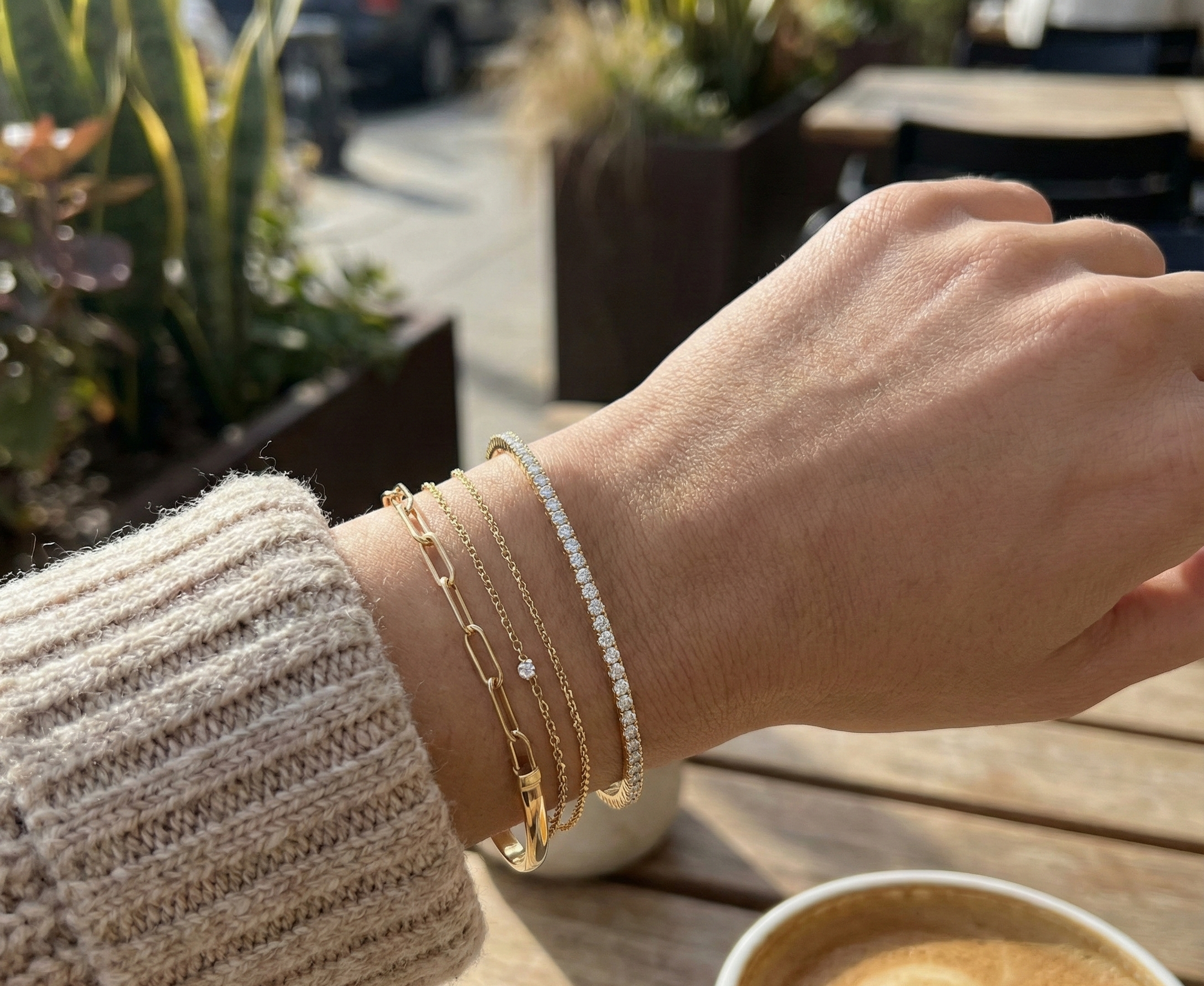 Hand wearing gold bracelets with a cup of coffee on a wooden table outdoors.